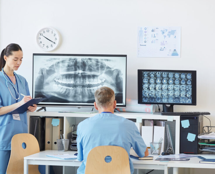 Back view at mature doctor sitting at desk in office of modern dental clinic and talking to nurse taking notes on clipboard, copy space